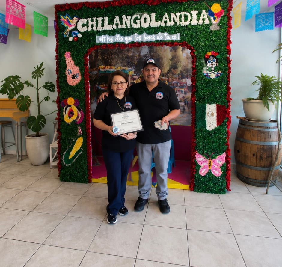 The two owners of Chilangolandia standing in front of the floral CHILANGOLANDIA photo wall, holding a certificate.